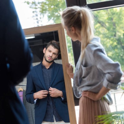woman grasping a full length mirror while man looks in the mirror trying on a sports coat